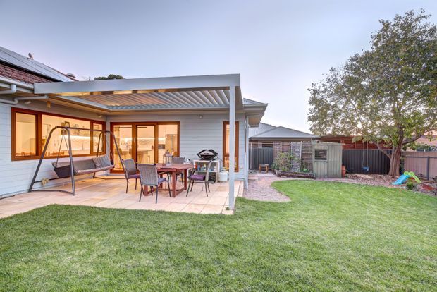 A suburban residential backyard with a covered patio extension attached to the house. The patio has a translucent pergola-style roof, pale horizontal exterior cladding, timber-framed glazed doors and windows, outdoor dining furniture, a barbecue, and a hanging swing chair. A paved path leads through a lawn toward a fenced garden area and shed.