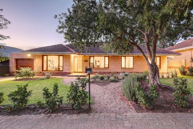 Single-storey brick suburban house with a low-pitched tiled hip roof, warm-lit windows and entry, and a landscaped front garden featuring gravel beds, shrubs, lawn, and a mature tree. A paved pedestrian path runs along the foreground.