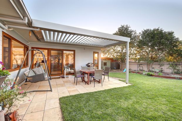 A landscaped residential backyard with a tiled patio adjoining a single-storey home. The patio includes an outdoor dining setting, hanging chair, and a large white pergola with a louvred roof structure. Timber-framed glazed doors and windows open onto the patio, with lawn, garden beds, and a boundary fence beyond.
