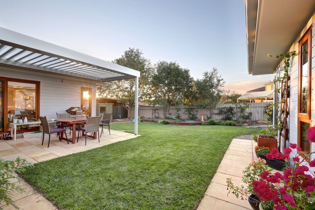 A landscaped backyard courtyard with a broad lawn, paved path, and covered outdoor dining area beneath a white pergola. The space includes an outdoor dining table and chairs, barbecue cabinetry, garden beds, timber-framed glazing, and flowering plants along the house.
