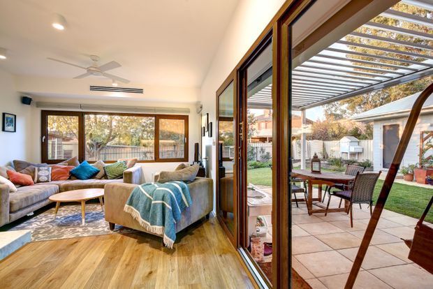 A bright, open-plan living room with timber flooring, large windows and extensive folding glass doors opening onto a paved outdoor patio and garden. The room contains upholstered sofas, a coffee table, patterned rug, ceiling fan and recessed lighting. The covered patio has outdoor dining furniture and a slatted pergola structure, creating a strong indoor–outdoor connection.
