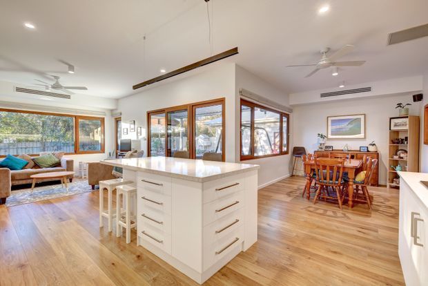 Bright open-plan residential interior combining a white kitchen with a central island, adjoining dining area, and living room. Warm timber flooring, extensive timber-framed glazing, white cabinetry, timber dining furniture, ceiling fans, and linear pendant lighting create an airy, contemporary family space.