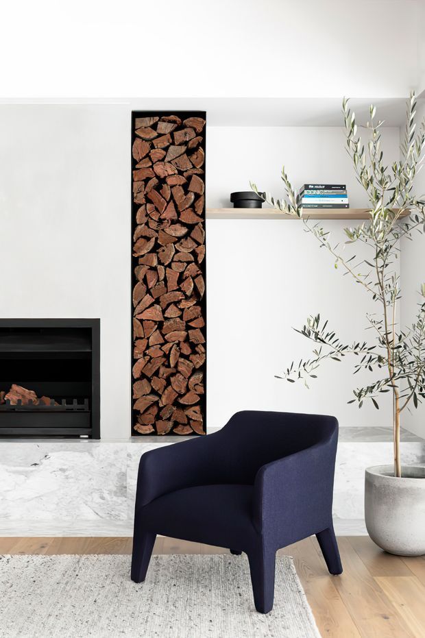 A refined living room interior featuring a minimalist black fireplace with an integrated vertical firewood storage niche, a white marble hearth, and a simple floating timber shelf. A dark navy upholstered armchair sits on a pale textured rug over timber flooring, while a potted olive tree adds an organic element. The space is bright, clean, and contemporary with strong black, white, timber, and navy contrasts.