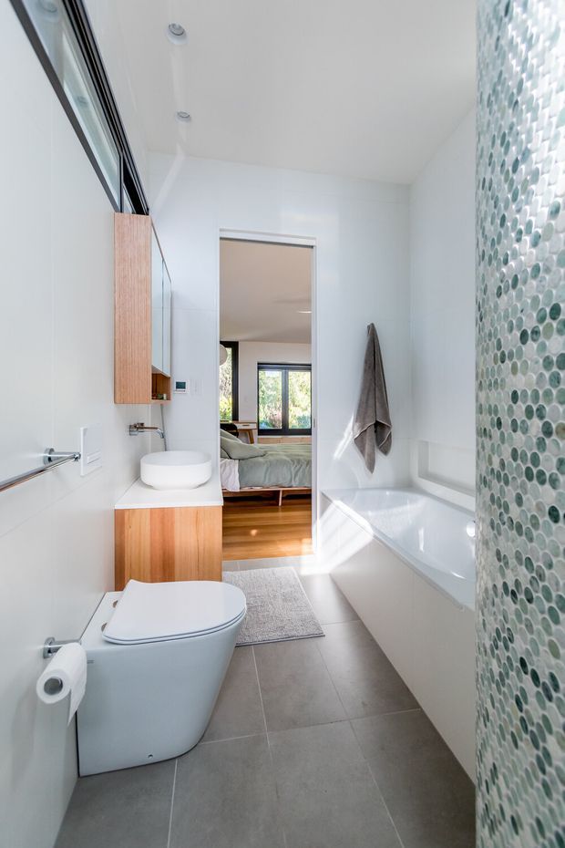 Bright contemporary bathroom with a white bathtub, wall-mounted toilet, round vessel basin, and warm timber vanity cabinetry. A green and white pebble mosaic tile wall forms a feature beside the tub, while grey large-format floor tiles and white walls create a clean, modern finish. The bathroom opens directly to a bedroom visible through the doorway.