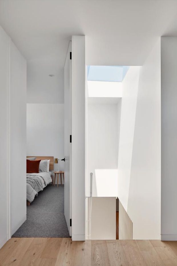 Bright minimalist upper-level hallway or landing overlooking a stairwell, with white walls and ceiling, pale timber flooring, a skylight, and a white balustrade. An open doorway reveals a bedroom with a bed and grey carpet. The space is clean and contemporary with strong natural light.