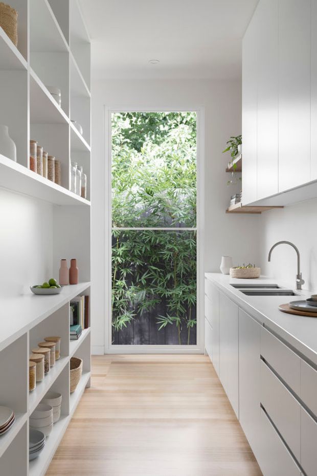 Bright, narrow galley kitchen with white full-height cabinetry, open white shelving, a pale stone benchtop, integrated sink and timber flooring. A tall glazed door or window at the rear looks onto dense greenery, bringing natural light into the minimalist space.
