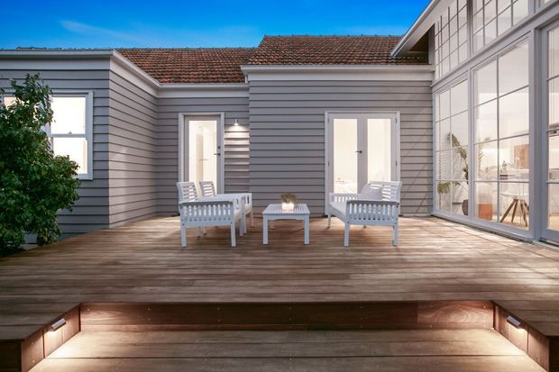 A bright courtyard-style timber deck adjoining a grey weatherboard house. White outdoor lounge furniture and a small table sit on the deck, with large glazed doors and windows opening to an interior room. The house has a low-pitched terracotta tiled roof and a raised deck with integrated step lighting.