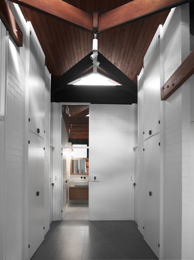A narrow, high-ceilinged interior passage lined with tall white built-in doors and storage cabinetry. The space features white painted brick walls, dark grey floor tiles, exposed dark timber beams, and a timber-lined vaulted ceiling with a triangular skylight. A bathroom or vanity area is visible at the end of the corridor.