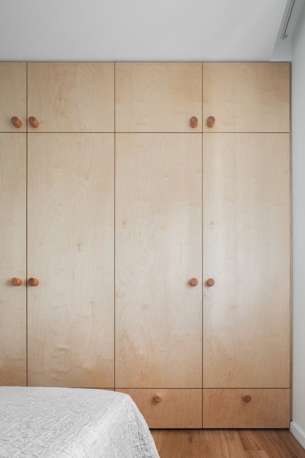 A close-up view of a built-in pale timber wardrobe occupying most of the wall in a bedroom. The cabinetry has tall flat-panel doors and lower drawers with small rounded wooden knobs, set against white walls and a timber floor. The corner of a bed with a light quilt is visible in the foreground.