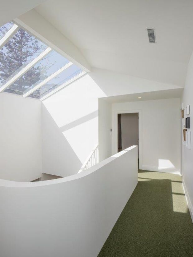 Bright minimalist upper-level hallway or landing with white walls, a curved half-height balustrade, green carpet, and a large sloping skylight flooding the space with natural light. A stairwell is visible beyond the curved wall, with a doorway and wall-mounted controls along the corridor.
