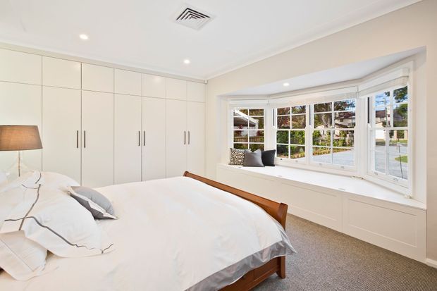 Bright, calm bedroom with a large timber-framed bed, white bedding, full-height built-in wardrobes, and a broad bay-style window with built-in cabinetry below. The room has light neutral finishes, recessed ceiling lights, carpet flooring, and a simple upholstered table lamp.
