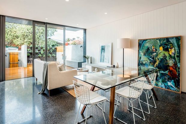 Bright open-plan modern living and dining area with polished dark terrazzo flooring, white vertical wall panelling, extensive floor-to-ceiling glazing, and a glass dining table with wire-frame chairs. A light upholstered lounge chair sits beside a low media console, while a large colourful artwork provides a bold accent. The glazing opens toward a landscaped outdoor courtyard or garden.