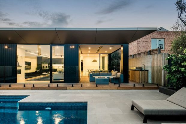 Contemporary single-storey home viewed from a paved pool terrace at dusk. The dark-clad pavilion has broad floor-to-ceiling glazing and an open living area, with a warm, light-filled interior visible through the windows. A pale brick adjoining structure, timber fencing, landscaped planting and a rectangular swimming pool complete the setting.