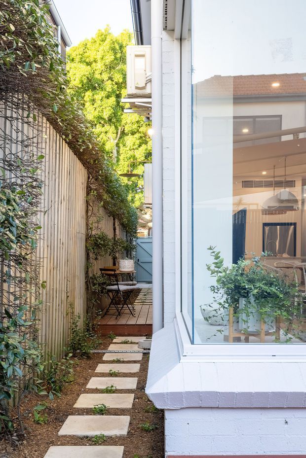 A narrow landscaped side passage beside a white-painted brick house. Stepping stones lead through a planted garden toward a small timber-deck seating nook with café chairs and a table. A tall timber boundary fence is covered with climbing greenery, while a large corner window provides views into a bright contemporary interior with pendant lighting and dining furniture.