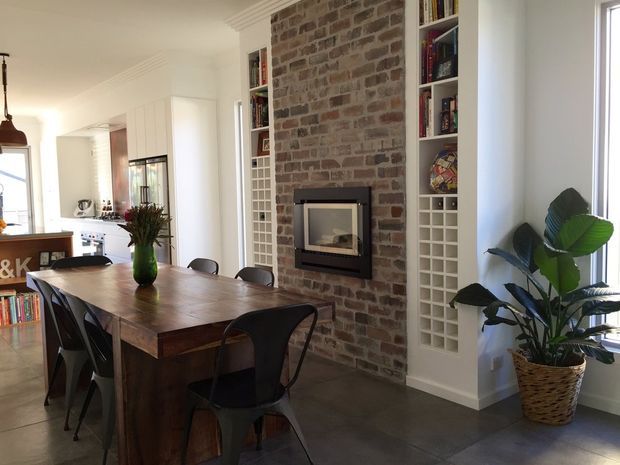 Open-plan dining area with a large dark timber dining table and black chairs, adjacent to a bright white kitchen. A prominent exposed brick fireplace wall with built-in white shelving and wine storage forms the main feature, with dark tiled flooring and abundant natural light.