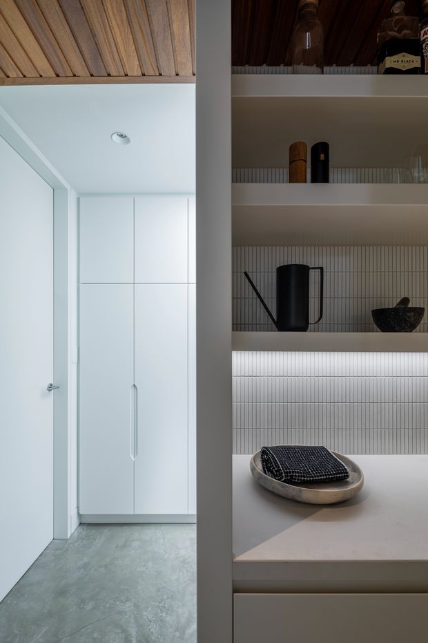 A refined built-in storage and display wall viewed beside a minimalist hallway or dressing area. The shelving features a vertically ribbed light neutral backsplash, integrated warm LED lighting, pale solid-looking benchtop and cabinetry, and carefully arranged dark decorative objects. Adjacent white full-height cupboards, a flush white door, grey polished concrete-style flooring, and a timber-lined ceiling create a restrained contemporary interior.