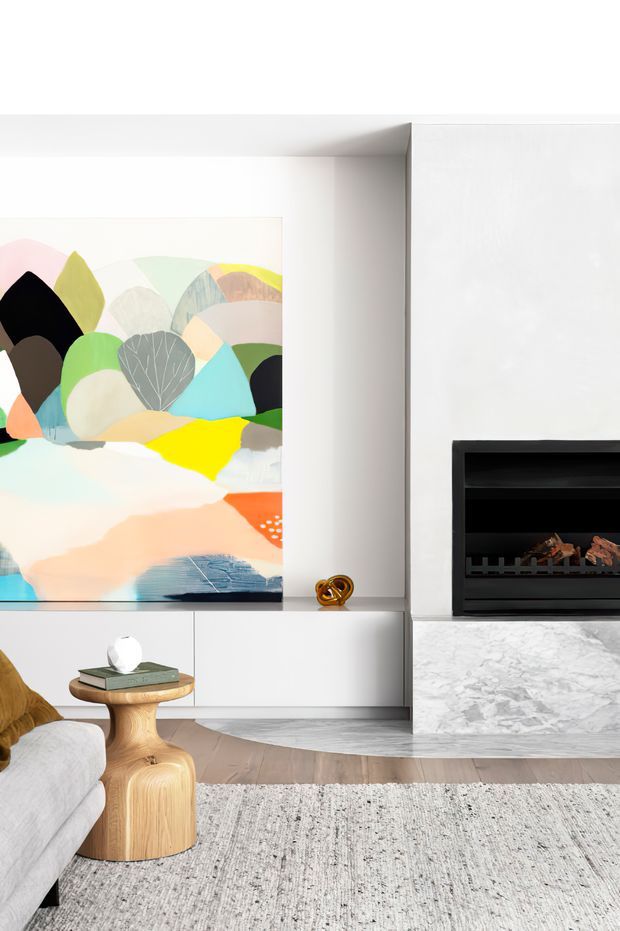 A bright, refined living room with a contemporary fireplace wall, white built-in cabinetry, a large colourful abstract artwork, pale upholstered seating, a sculptural timber side table, and a light textured rug. The fireplace is framed by a grey-white marble surround and hearth, while timber flooring adds warmth.