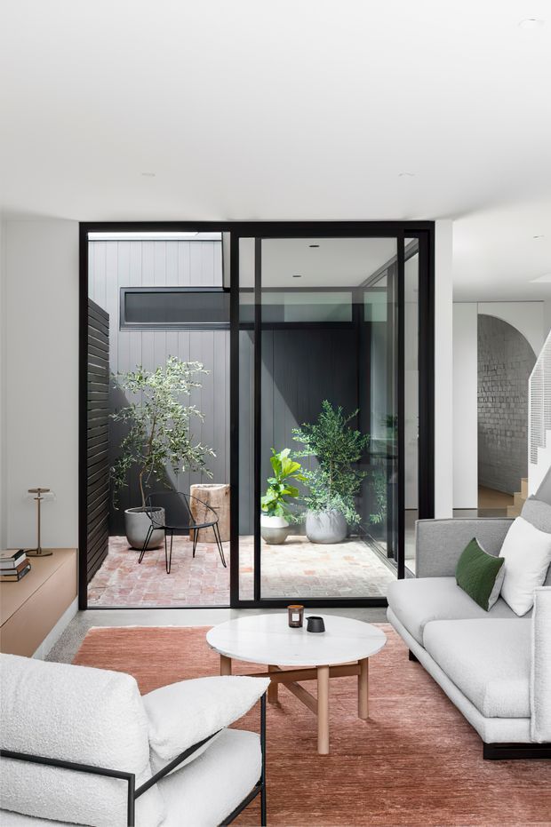 A bright contemporary living room with a large light-coloured sectional sofa, sculptural lounge chair, round white coffee table, and muted terracotta rug. Black-framed floor-to-ceiling glazing opens visually onto a small internal courtyard with potted plants and brick paving. White walls and ceiling, dark timber accents, and a glimpse of an arched masonry passage create a clean, refined interior.