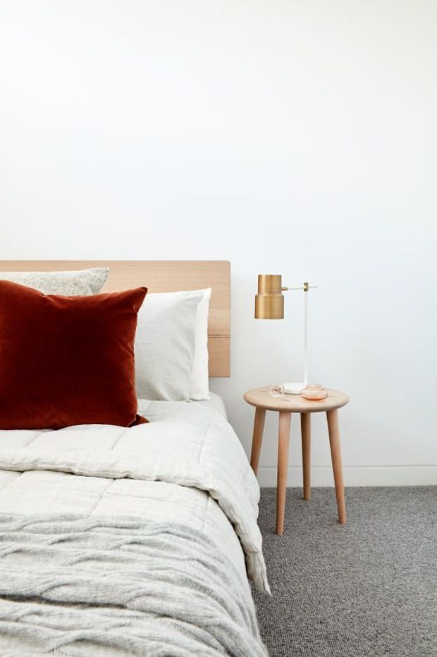 A bright, minimalist bedroom corner featuring a low light-timber bedhead, white layered bedding, a deep rust-red velvet cushion, and a small round timber bedside table. A slim brass/gold wall-mounted reading lamp is positioned above the table, with grey carpet flooring and plain white walls.