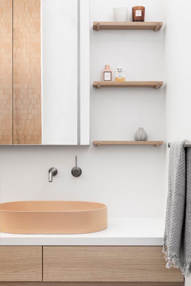 A bright, minimalist bathroom vanity vignette featuring a peach-toned vessel basin on a white countertop, brushed metal wall-mounted tapware, light timber floating vanity cabinetry, and three matching timber display shelves with toiletries and ceramics. A large mirrored cabinet with a vertical timber mosaic panel occupies the left side, while a textured grey towel hangs at right.