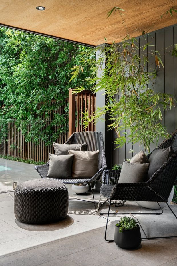 A stylish covered outdoor patio or terrace furnished with two black woven lounge chairs, neutral cushions, and a matching round ottoman. Large pale grey paving tiles, a dark vertical-panel wall, timber ceiling, glass balustrade, and dense bamboo and greenery create a private, contemporary garden setting.
