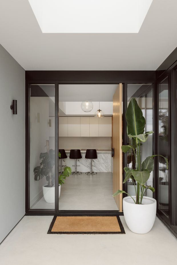A refined entryway viewed from a light concrete-finished threshold, looking through dark-framed glazed doors into a contemporary kitchen and dining area. The entry features a large rectangular skylight, dark timber or metal-framed glazing, a timber pivot door, a tan and black doormat, and tall potted tropical plants. Beyond the doorway are pale built-in kitchen cabinets, a marble-look island or splashback, and dark bar stools.