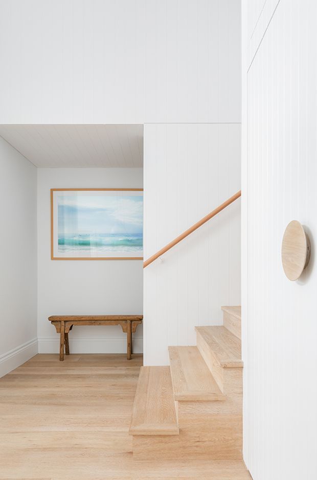 Bright minimalist residential stairway and landing with pale timber flooring and open-riser stairs, white vertical wall panelling, a simple timber handrail, and a narrow console table beneath a framed coastal artwork. The space is airy and restrained, with light natural materials and clean architectural lines.
