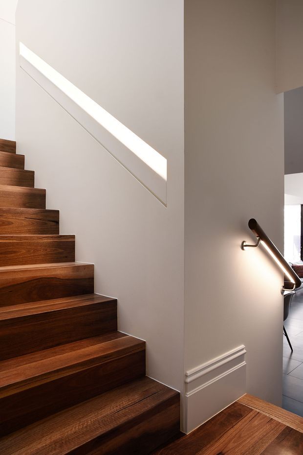 Interior view of a refined stairway with richly stained dark timber treads and matching timber flooring. The staircase is enclosed by smooth warm-grey painted walls, with a slim integrated handrail and a sculptural wall-mounted light. Decorative linear wall mouldings add a traditional detail, while daylight enters through a narrow angular opening.