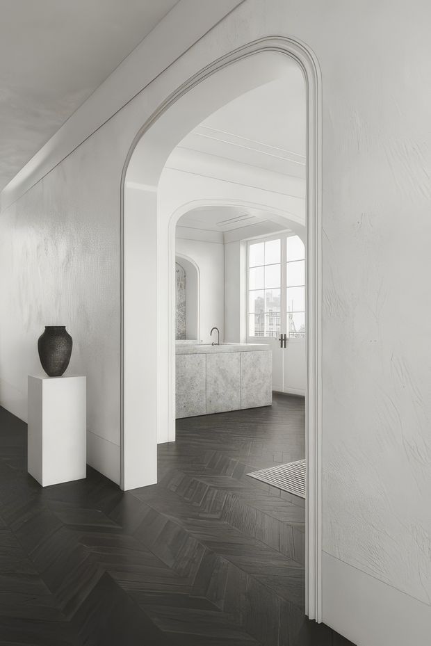A refined monochromatic interior view through a tall arched doorway into a bright kitchen or living space. The foreground has dark herringbone timber flooring, textured white walls, sculptural trim around the arch, and a white pedestal displaying a dark ceramic vase. Beyond the doorway is a pale stone island with a black tap, white paneled ceiling, large multi-pane glazing, and another arched opening. The overall composition is calm, contemporary, and highly tonal.