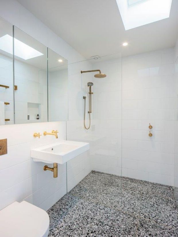 Bright contemporary bathroom with white rectangular wall tiles, a wall-mounted white basin, large mirrored wall panels, brass/gold tapware and shower fittings, a frameless glass shower area, a terrazzo-style speckled floor, and a skylight.