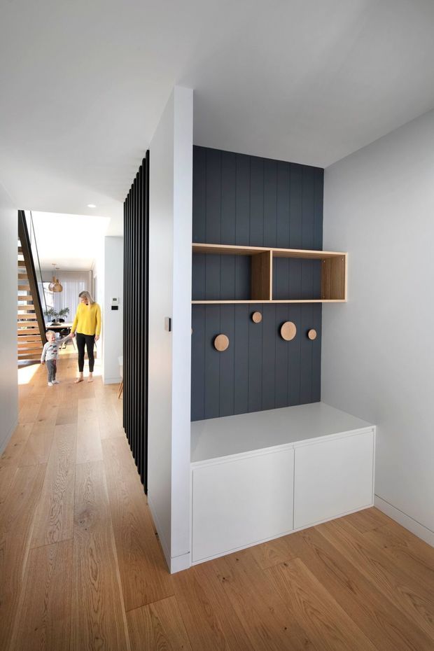A bright contemporary hallway and entry zone with wide light-oak timber flooring, white walls and ceiling, and a built-in storage/display unit. The feature joinery combines a dark charcoal vertical-panelled wall with open mid-wood shelving, a white low cabinet, and small round timber pegs. A black vertical slatted screen separates the hallway from the adjacent living area, where a timber stair and pendant light are visible.