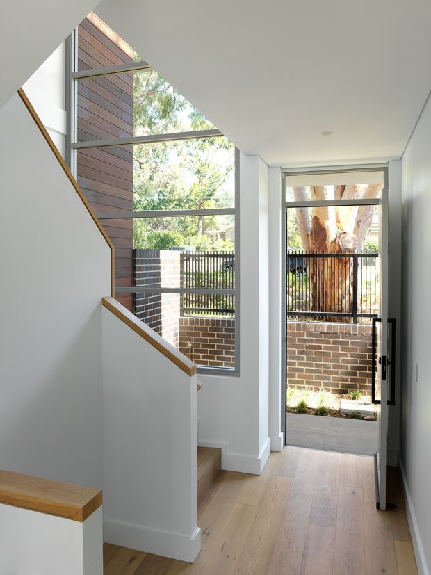 Bright contemporary entry hall with a timber stair and white painted balustrade, pale timber flooring, full-height glazed windows, and a glazed external door opening to a small brick-walled garden or courtyard. Black metal window and door frames contrast with the white walls, while mature trees are visible outside.