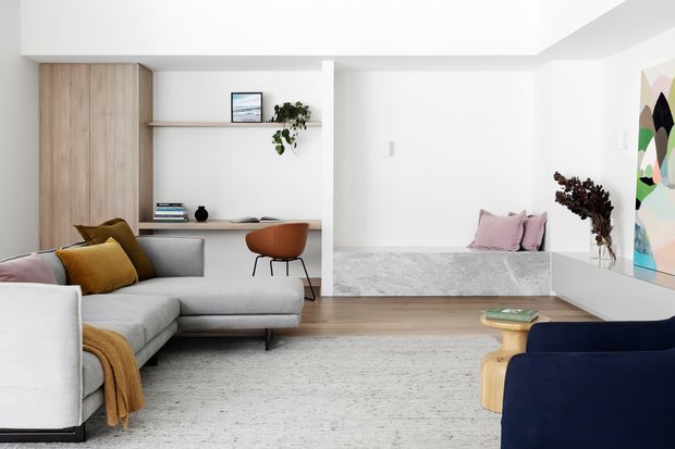 Bright, refined contemporary living room with a pale grey sectional sofa, navy lounge chair, textured light rug, timber flooring, white walls, and a built-in timber-lined study nook with floating shelf and desk. A raised marble-look platform with cushions and colourful artwork adds visual interest.