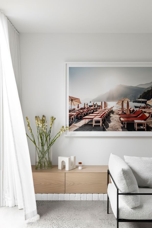 Bright, minimalist living room with white walls, sheer floor-to-ceiling curtains, a light timber low sideboard, white textured armchair, and a large framed artwork depicting a lakeside terrace. A glass vase with tall branches adds a natural accent, while a pale terrazzo-like floor and white-painted brick base provide subtle texture.
