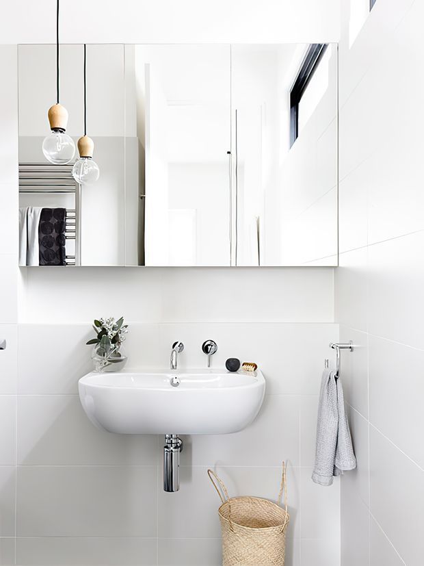 Bright, minimalist bathroom vanity with a wall-mounted white basin, chrome tapware, large mirrored cabinet, white tiled walls, pendant lights, a heated towel rail, and a woven basket. The mirror reflects the clean, contemporary bathroom interior.