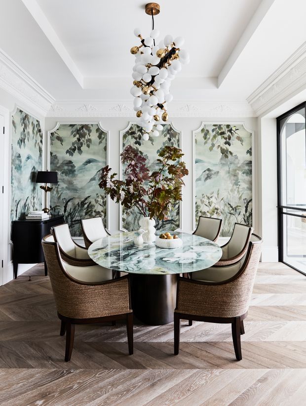 Elegant formal dining room with a round marble-topped dining table, upholstered dining chairs, ornate white wall mouldings, botanical mural panels, dark timber herringbone flooring, and a dramatic sculptural brass-and-white-globe chandelier. Large glazed doors bring in abundant natural light.