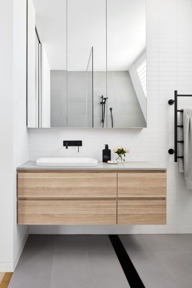 Bright contemporary bathroom featuring a wall-mounted light timber vanity with integrated drawers, a white vessel basin, large mirrored cabinetry, white horizontal wall tiles, grey floor tiles, matte black tapware and shower fittings, and a black ladder-style towel rail. A linear black floor drain runs toward the shower area.