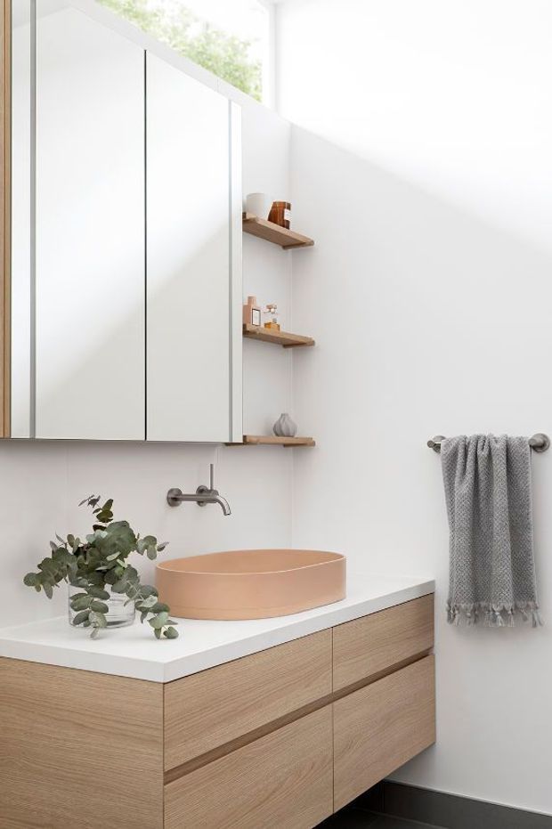 A bright, minimalist bathroom featuring a floating timber vanity with a white benchtop, a round vessel basin, wall-mounted brushed metal tapware, mirrored cabinetry, open timber shelves, and a grey towel rail. Soft daylight and restrained natural materials create a clean, warm contemporary interior.