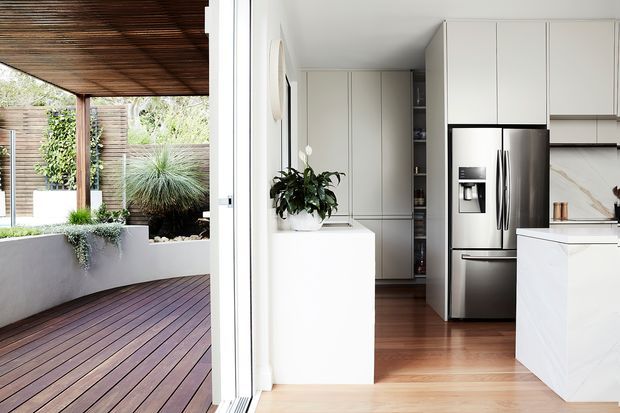 Bright contemporary kitchen opening directly onto a covered timber deck and landscaped courtyard. The kitchen features white floor-to-ceiling cabinetry, a white island, stainless-steel French-door refrigerator, pale timber flooring, and a marble-look splashback. The adjacent outdoor area has timber decking, a timber slatted ceiling, planted built-in garden beds, and a timber privacy screen.