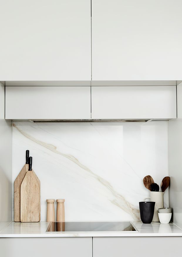 A close-up of a minimalist contemporary kitchen featuring white handleless cabinetry, a large white marble-look splashback with subtle veining, and a flush-mounted stainless steel cooktop integrated into the benchtop. Wooden chopping boards, timber-toned canisters, and dark utensils provide restrained natural and dark accents.