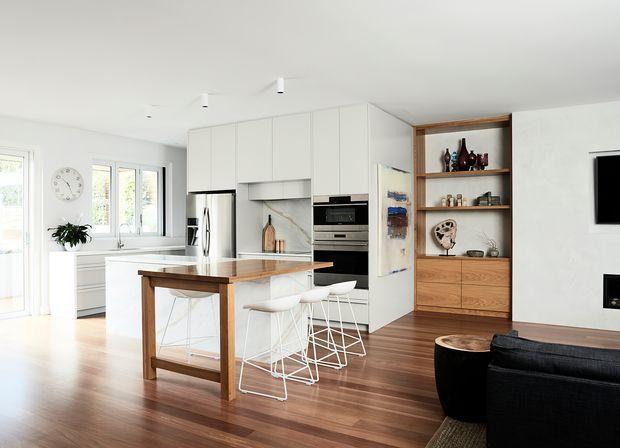 Bright open-plan contemporary kitchen and living area with white handleless cabinetry, integrated stainless-steel appliances, a timber-topped island with white bar stools, warm timber flooring, and built-in timber shelving displaying decorative objects. Large windows provide abundant natural light, while a dark sofa and wall-mounted television occupy the living zone.