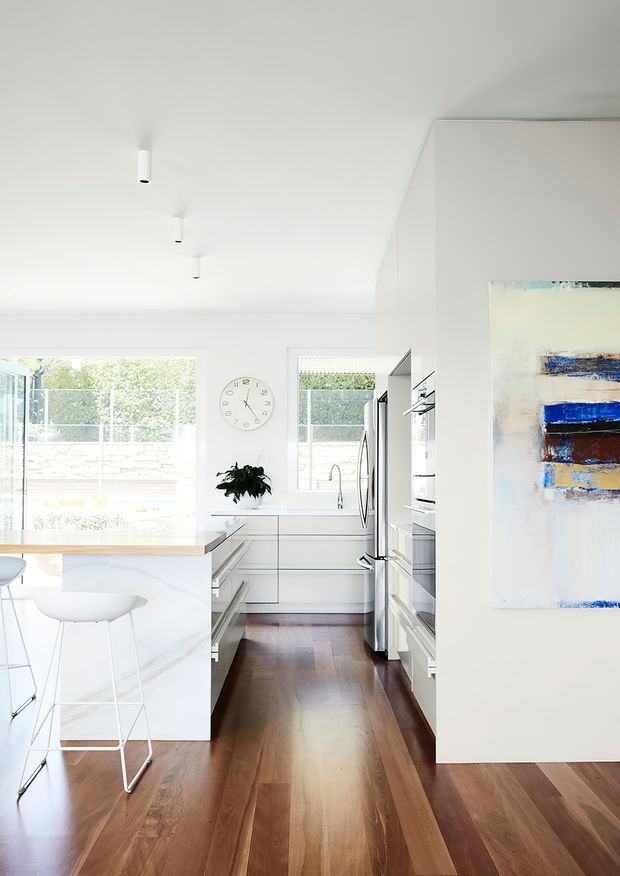 Bright, modern open-plan kitchen with white cabinetry, a white marble-look island and waterfall edge, stainless-steel appliances, dark timber flooring, and large windows overlooking an outdoor area. Minimal white walls and ceiling are complemented by a colourful abstract artwork and simple bar seating.