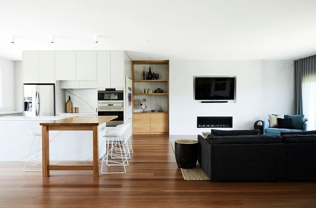 Open-plan contemporary interior combining a white kitchen with island bench and bar stools, a timber dining table, and a living area with a dark sectional sofa. Warm timber flooring and a built-in timber display cabinet contrast with the white walls and cabinetry. A wall-mounted television and linear fireplace form the living-room focal point, with large curtained glazing at right.