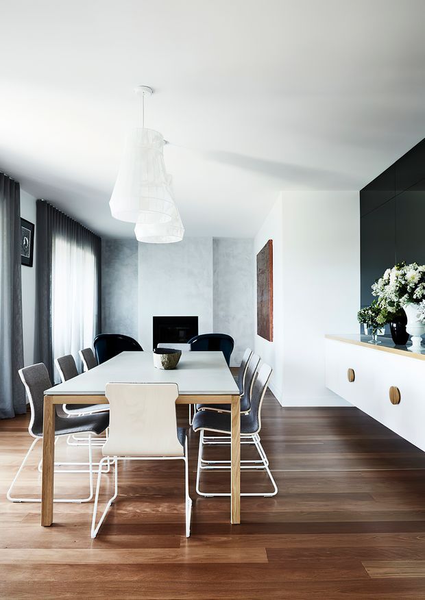 Bright contemporary dining area with a long pale dining table surrounded by mixed modern chairs, set over dark timber flooring. The room features floor-to-ceiling sheer and dark curtains, a white sculptural pendant light, a minimalist black fireplace, white walls, and a sleek black built-in cabinetry unit with a pale benchtop and decorative floral arrangement.