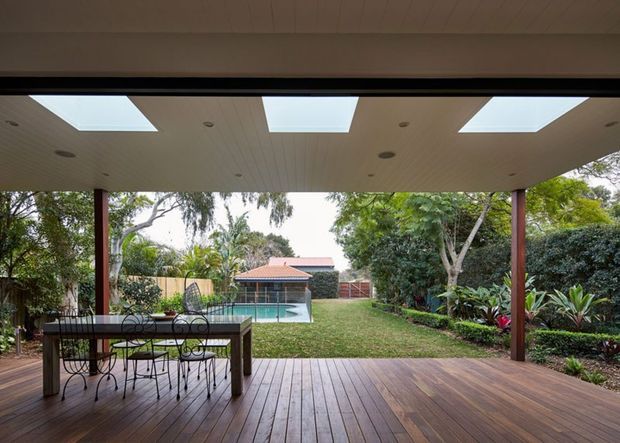 Covered outdoor entertaining deck opening onto a landscaped backyard, with a timber dining table and chairs, broad timber decking, slender posts, and a soffit incorporating three skylights and recessed lights. A swimming pool, lawn, hedges, and garden planting are visible beyond.