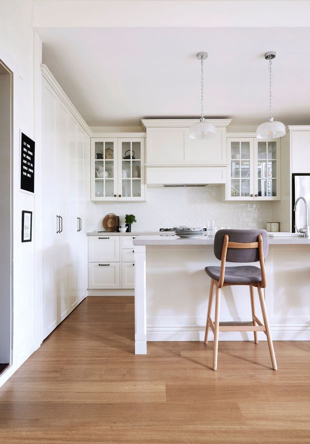 Bright, refined white kitchen with full-height panelled cabinetry, glass-front upper cabinets, a central island with seating, white subway-tile splashback, timber flooring, and pendant lights. The space has a classic yet contemporary residential fit-out.