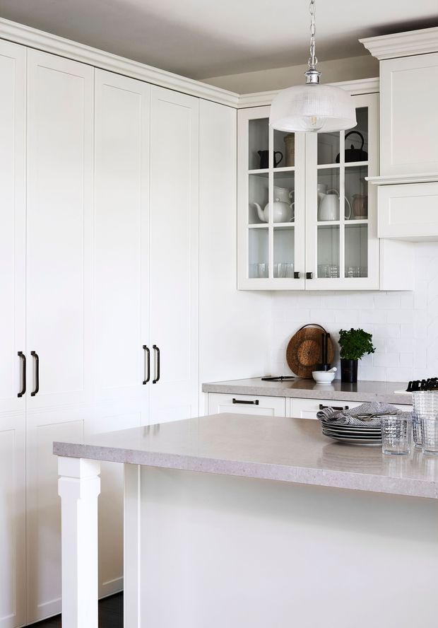 Bright, predominantly white traditional kitchen with extensive recessed-panel cabinetry, a large peninsula with a pale stone benchtop, glass-front display cabinets, white subway-tile splashback, and classic moulded cornices. Black hardware, a pendant light, crockery, and greenery provide restrained accents.