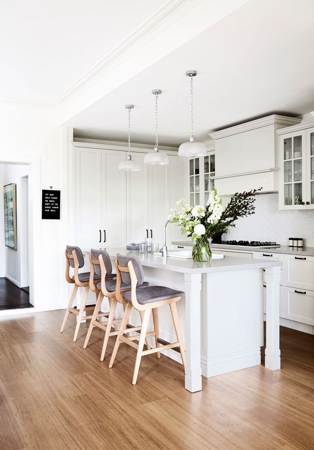 Bright, spacious white kitchen with classic panelled cabinetry, glass-front upper cupboards, a large central island with seating for four, pendant lights, white benchtops and a timber floor. The room has refined traditional detailing with warm wood and upholstered bar stools.