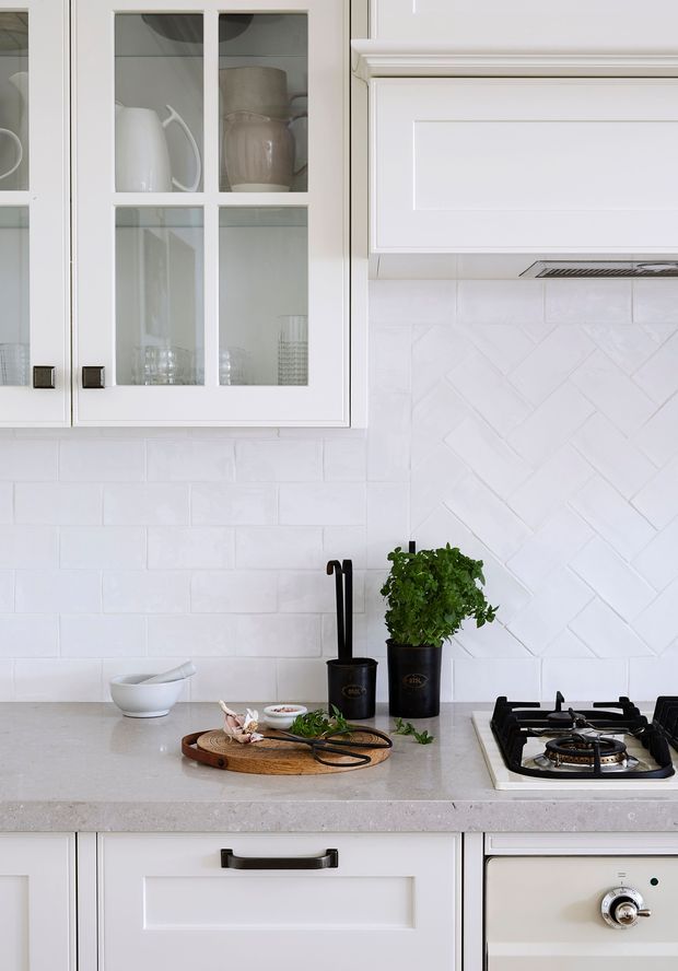 Close-up view of a bright, refined kitchen featuring white shaker-style cabinetry with glass-front upper doors, white subway tile and herringbone tile splashbacks, pale engineered-stone benchtops, dark hardware, and a gas cooktop. Small potted herbs and kitchenware add natural detail.