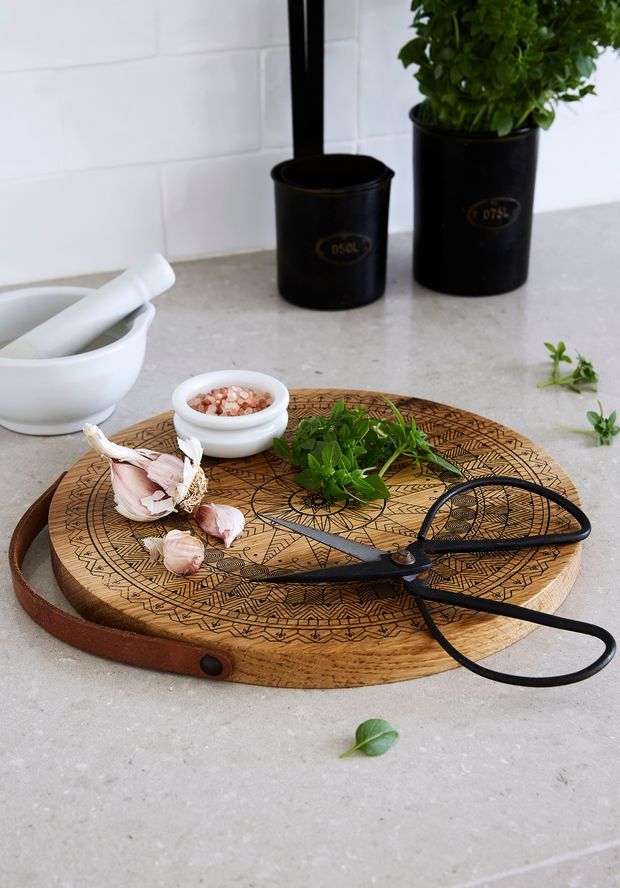 Still-life kitchen scene featuring a round engraved wooden serving board with a leather handle, black kitchen scissors, garlic, fresh herbs, and a small bowl of coarse salt. The board sits on a light stone countertop against a white tiled backsplash, with a white mortar and pestle and black potted plants in the background.